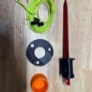 Puede incluir: Componentes de equipo de camping: una estaca roja con mango negro, una pieza circular negra, una tapa naranja y una bobina de cuerda verde neón con cierres negros. Dispuestos sobre una superficie de madera.