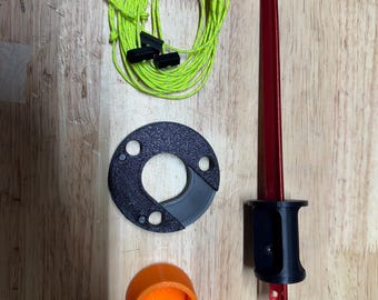 Puede incluir: Componentes de equipo de camping: una estaca roja con mango negro, una pieza circular negra, una tapa naranja y una bobina de cuerda verde neón con cierres negros. Dispuestos sobre una superficie de madera.