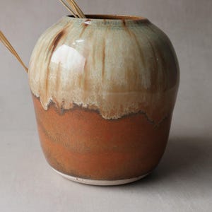 May include: A glazed ceramic vase with a bulbous shape and a wide mouth. The vase features a two-tone glaze, with a cream-coloured top and a reddish-brown base. A dried plant stem is placed inside the vase.