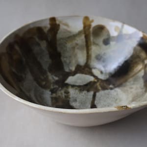 May include: A ceramic bowl with a cream exterior and a glazed interior featuring abstract brown and grey patterns. The bowl has a wide, shallow design, suitable for serving or display.