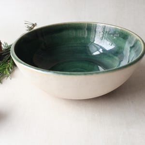 May include: A ceramic bowl with a cream-coloured exterior and a deep green interior. The bowl has a wide, open shape. Decorative elements include pine needles and a dried seed pod.