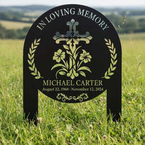 May include: Black metal memorial marker with the text "IN LOVING MEMORY" and the name "MICHAEL CARTER" with birth and death dates. The design includes a cross, floral elements, and laurel wreaths. The marker is placed in a grassy field.