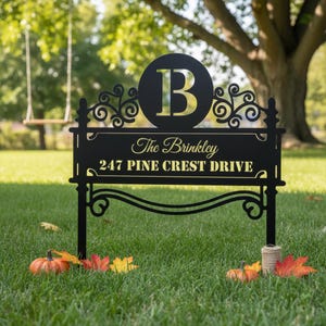 May include: Black metal yard sign with the letter "B" in a circle, the family name "The Brinkley", and the address "247 PINE CREST DRIVE" in gold lettering. The sign is in a grassy yard with fall leaves and pumpkins.
