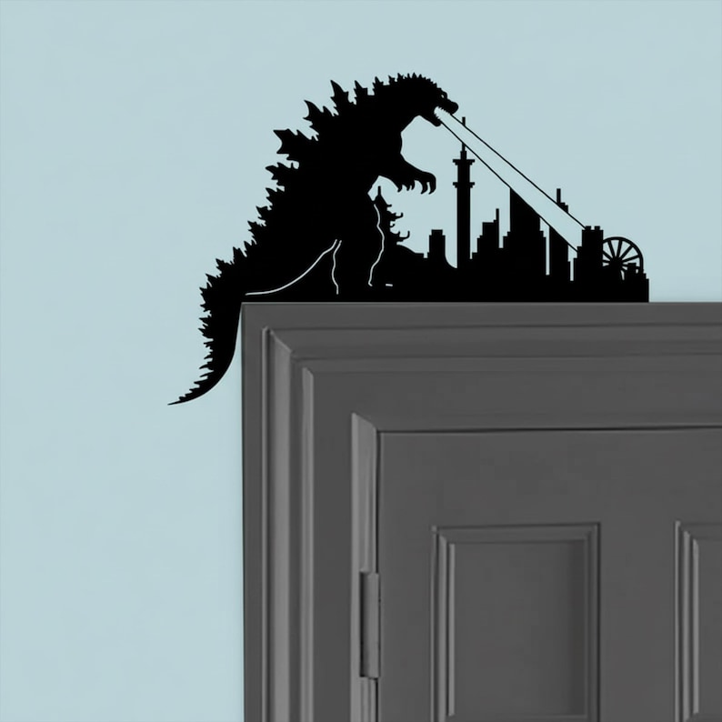 Puede incluir: Una silueta negra de Godzilla destruyendo un horizonte urbano. El monstruo se representa con la boca abierta, emitiendo un rayo de energ&iacute;a hacia los edificios. El dise&ntilde;o est&aacute; colocado en un marco de puerta.