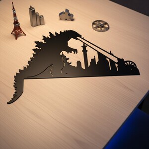 Puede incluir: Silueta de metal negro de Godzilla destruyendo una ciudad, con un rayo de energ&iacute;a. Edificios de metal en miniatura y una r&eacute;plica roja de la Torre Eiffel sobre una superficie de madera clara.