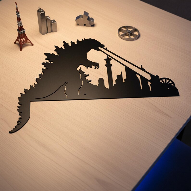 Puede incluir: Silueta de metal negro de Godzilla destruyendo una ciudad, con un rayo de energ&iacute;a. Edificios de metal en miniatura y una r&eacute;plica roja de la Torre Eiffel sobre una superficie de madera clara.