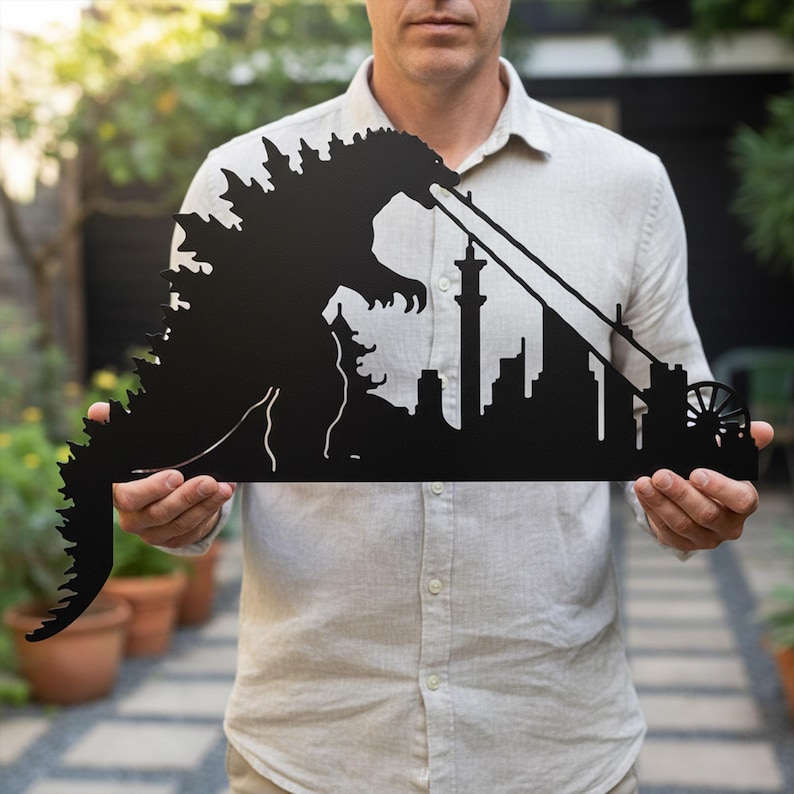 Puede incluir: Arte de pared de metal negro que representa a Godzilla destruyendo una ciudad. La silueta muestra al ic&oacute;nico monstruo lanzando fuego a un paisaje urbano con una torre alta y una noria. La obra de arte est&aacute; sostenida por una persona.