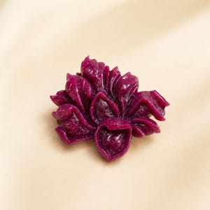Op de afbeelding: Een diep magenta broche in de vorm van een bloeiende bloem met meerdere gelaagde, gebogen bloemblaadjes. Het oppervlak van dit bloemvormige accessoire heeft een glanzende, gladde afwerking met ingewikkelde, gegoten details die diepte creëren.
