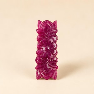 May include: A rectangular, deep magenta-coloured gemstone carved with a symmetrical floral pattern. The surface features intricate, raised petal and leaf motifs that run vertically along the length of the stone, creating a textured, relief-style design.