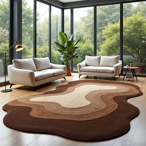 May include: A large, abstract area rug with a layered design in shades of brown, tan, and cream. The rug is placed on a hardwood floor in a room with two sofas and large windows overlooking a green landscape.