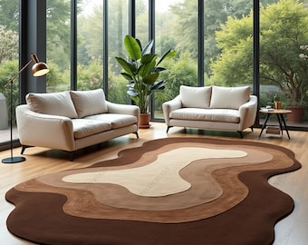 Puede incluir: Una alfombra grande y abstracta con un diseño en capas en tonos marrones, beige y crema. La alfombra está colocada sobre un suelo de madera en una habitación con dos sofás y grandes ventanales con vistas a un paisaje verde.