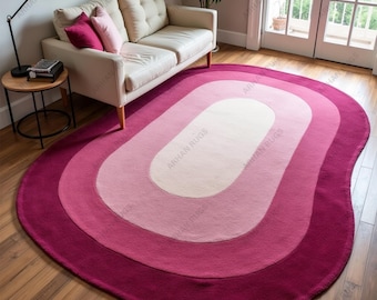 May include: A large, oval-shaped rug with a layered design in shades of pink and magenta. The rug features a central off-white oval, surrounded by layers of light pink, medium pink, and a deep magenta border. The rug is placed on a hardwood floor.