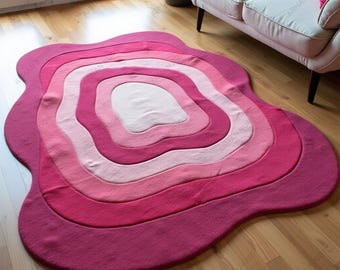 May include: A layered, abstract rug in shades of pink and magenta. The rug features a unique, organic shape with concentric rings of color, creating a gradient effect. The rug is placed on a hardwood floor, with a sofa and window in the background.