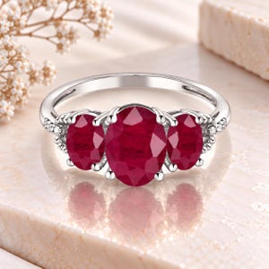 Puede incluir: Un anillo de tono plateado con tres piedras preciosas de color rojo intenso de corte ovalado dispuestas horizontalmente. La piedra central es más grande que las dos laterales. La banda está decorada con pequeñas piedras claras redondas.