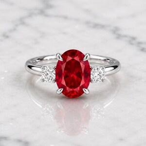 Op de afbeelding: Een zilverkleurige ring met een grote, ovaal geslepen, levendig rode edelsteen in het midden, vastgehouden door vier pootjes. De ringband is aan weerszijden geaccentueerd met een kleinere, rond geslepen, helderwitte edelsteen.