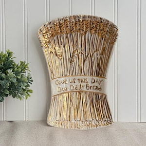 May include: A white ceramic serving tray shaped like a sheaf of wheat with gold-painted details. The tray features embossed wheat stalks and the text "GIVE US THIS DAY Our Daily Bread" in gold lettering across the center of the wheat bundle.