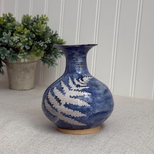 May include: A blue ceramic vase with a white fern design. The vase has a rounded body and a narrow neck. It sits on a small, round, tan base. A green plant in a pot is in the background.