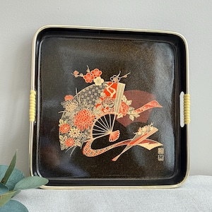 May include: Square black tray with gold trim and yellow handles. The tray features a decorative Japanese-style design with flowers, fans, and geometric patterns in red, orange, and gold. The tray is likely used for serving or display.
