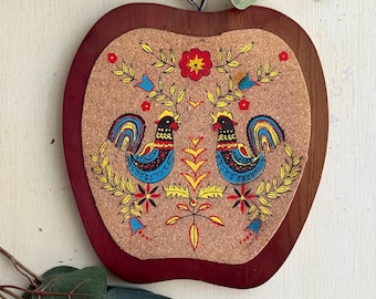 May include: Apple-shaped trivet with a dark red wooden frame and a cork center. The cork features a colorful folk art design of two roosters, flowers, and foliage in yellow, blue, and red. A black loop is attached for hanging.
