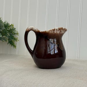May include: A small, brown ceramic pitcher with a handle. The top edge has a white and brown glaze. The pitcher is sitting on a neutral-colored surface, with a white-paneled wall in the background.