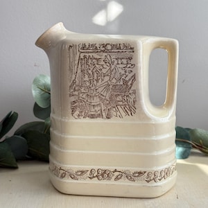 May include: Cream-colored ceramic pitcher with a brown illustration of a person working. The pitcher has a rectangular handle and horizontal ridges. A brown floral pattern decorates the base. The pitcher is a vintage style.