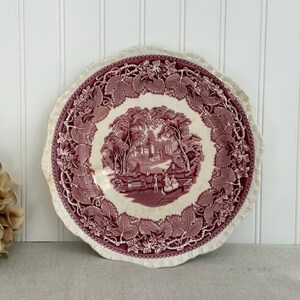 May include: A round ceramic plate with a scalloped edge featuring a deep maroon transferware pattern. The center depicts a landscape with a castle, trees, and figures, while the border is decorated with a dense pattern of grape leaves and vines.