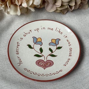 May include: A small, round, off-white ceramic plate with a red rim. The plate features a floral design with a heart and the text "Your secret is shut up in me like it was dead once."