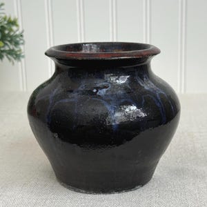 May include: A handcrafted, dark-glazed ceramic vase with a rounded body and a slightly flared rim. The vase has a glossy finish with subtle blue and red accents, showcasing a rustic aesthetic. This decorative piece is suitable for displaying flowers or as a standalone art object.
