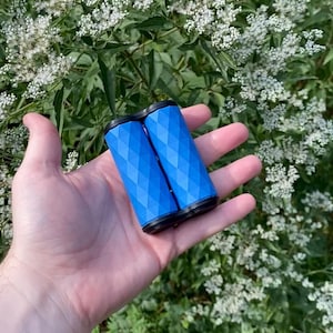 May include: A pair of blue bicycle handle grips with a diamond pattern, held in a hand against a backdrop of green foliage and white flowers. The grips have black ends.