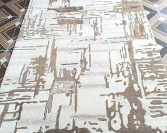 May include: Rectangular rug with an abstract design in beige and brown tones. The rug has a textured pattern with vertical and horizontal lines and shapes, creating a modern look. The rug is placed on a tiled floor.