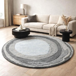 Könnte beinhalten: Ein runder Teppich mit einem geschichteten Design in Grau- und Hellblautönen. Der Teppich hat eine strukturierte Oberfläche und befindet sich in einem Wohnzimmer. Der Teppich trägt den Text "Haniya Rugs Weaver".