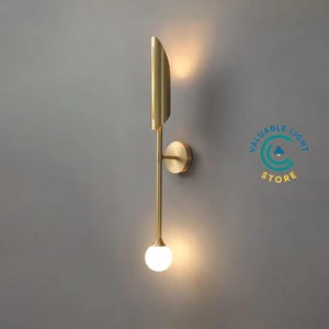 May include: A modern wall sconce with a brass finish. The sconce features a vertical design with a curved top, a circular base, and a spherical, frosted glass light at the bottom. The text "VALUABLE LIGHT STORE" is visible.