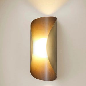 May include: A modern wall sconce with a warm, inviting glow. The fixture has a curved, metallic shade in a bronze tone, creating a soft, ambient light. The light source is a bright, central bulb, perfect for adding a touch of elegance to any space.
