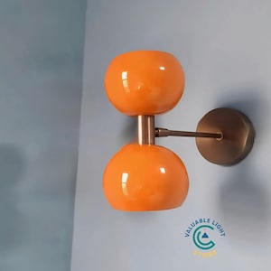 Puede incluir: Aplique de pared moderno con dos pantallas globulares naranjas. La lámpara tiene un acabado bronce y está montada en una pared azul claro. El diseño es contemporáneo, adecuado para la iluminación interior.