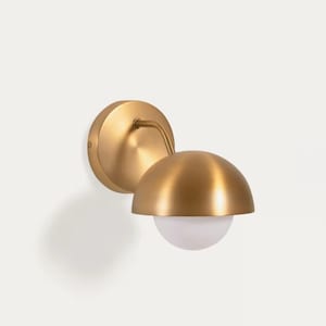 May include: A brass-colored wall sconce with a semi-spherical shade and a white, globe-shaped light. The sconce is mounted on a circular base and has a curved arm connecting the shade to the base. The fixture is designed for indoor use.