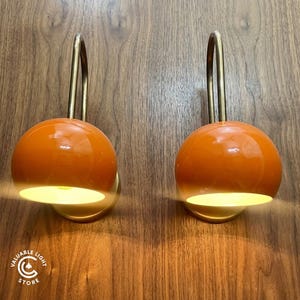May include: Two wall lamps featuring glossy orange dome-shaped shades attached to curved brass-toned metal arms. Each lamp emits a warm light from the underside of the dome. A circular logo with the text VALUABLE LIGHT STORE is in the corner.