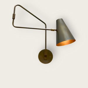 Könnte beinhalten: Eine Wandleuchte mit konischem Schirm und Messing-Finish. Die Lampe hat einen verstellbaren Arm und einen kreisförmigen Sockel. Die Innenseite des Schirms hat einen warmen, goldenen Farbton. Das Design ist modern und minimalistisch.