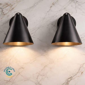 May include: Two black cone-shaped wall sconces with a gold interior. The sconces are mounted on a marble wall. The sconces have a modern design and emit a warm light. The sconces are a stylish addition to any home decor.