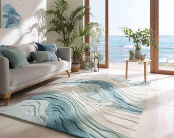 Puede incluir: Una alfombra grande con un diseño de olas en tonos azules y beige. La alfombra está colocada en un salón con un sofá gris, cojines y plantas. La habitación tiene una gran ventana con vistas al océano.