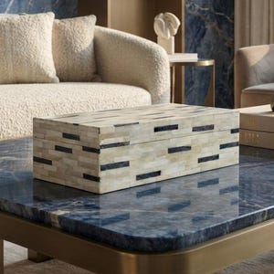 May include: A rectangular decorative box featuring a mosaic pattern of cream-colored and black rectangular inlays. The box has a smooth, flat lid and sits on a polished blue stone surface with a gold-toned metal frame and four sturdy legs.