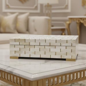 May include: A rectangular decorative box with a brick-like pattern in cream and gold tones. The box is placed on a marble-topped table with a gold-colored frame. The box has gold-colored feet.