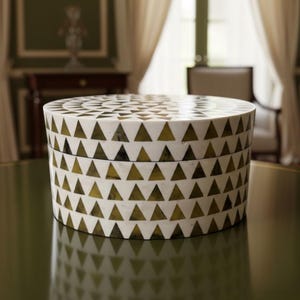 May include: A round, decorative box with a white background and a repeating pattern of green triangles. The box is sitting on a green surface, and the background includes curtains and furniture.