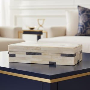 May include: Rectangular decorative box with a cream-colored mosaic pattern and dark gray accents. The box is placed on a dark blue table with a gold trim. The background features a white sofa and a gold-trimmed table.