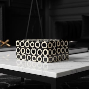 May include: A rectangular black and white decorative box with a repeating pattern of white circles on a black background. The box is on a white marble table with black legs. A gold-colored decorative object is visible in the background.