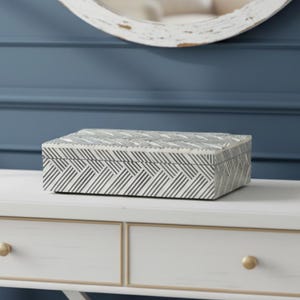 May include: A rectangular decorative box with a geometric black and white pattern. The box is placed on a white table with gold-colored drawer pulls. A white-framed mirror is visible in the background.