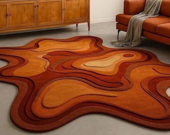 Op de afbeelding: Een onregelmatig gevormd vloerkleed met een gelaagd, topografisch ontwerp in verschillende tinten oranje, roest en crème. Het kleed heeft een getextureerd oppervlak met meerdere niveaus en gebogen, vloeiende lijnen.