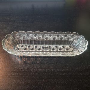 May include: An elongated, clear glass dish with a scalloped rim and a textured geometric pattern featuring stars and circles. The center of the base displays the embossed text AVON. The dish has a shallow, oval shape with vertical side walls.