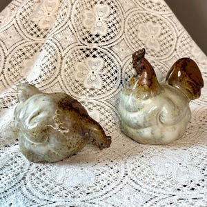 May include: Two ceramic chicken figurines. One is a hen, the other a rooster. The hen is light brown and cream-colored, with its head down. The rooster is brown and cream-colored, with a brown comb and tail feathers. The figurines are on a white lace tablecloth.