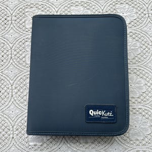 May include: A navy blue zippered binder with rounded corners. The binder has a small rectangular label that reads "QuicKutz" in white text. The background is a white lace tablecloth.
