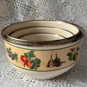 May include: Set of three nesting bowls with a cream-colored interior and a beige exterior band featuring fruit illustrations. The bowls have a metallic silver rim and a green border. The bowls are stacked on top of each other.